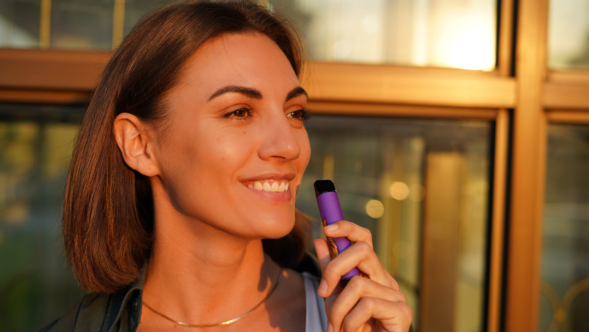 Lächelnde Frau, die bei Sonnenuntergang im Freien einen Vape Pen benutzt, um den Lebensstil der Reduzierung des Nikotinkonsums durch Dampfen darzustellen.