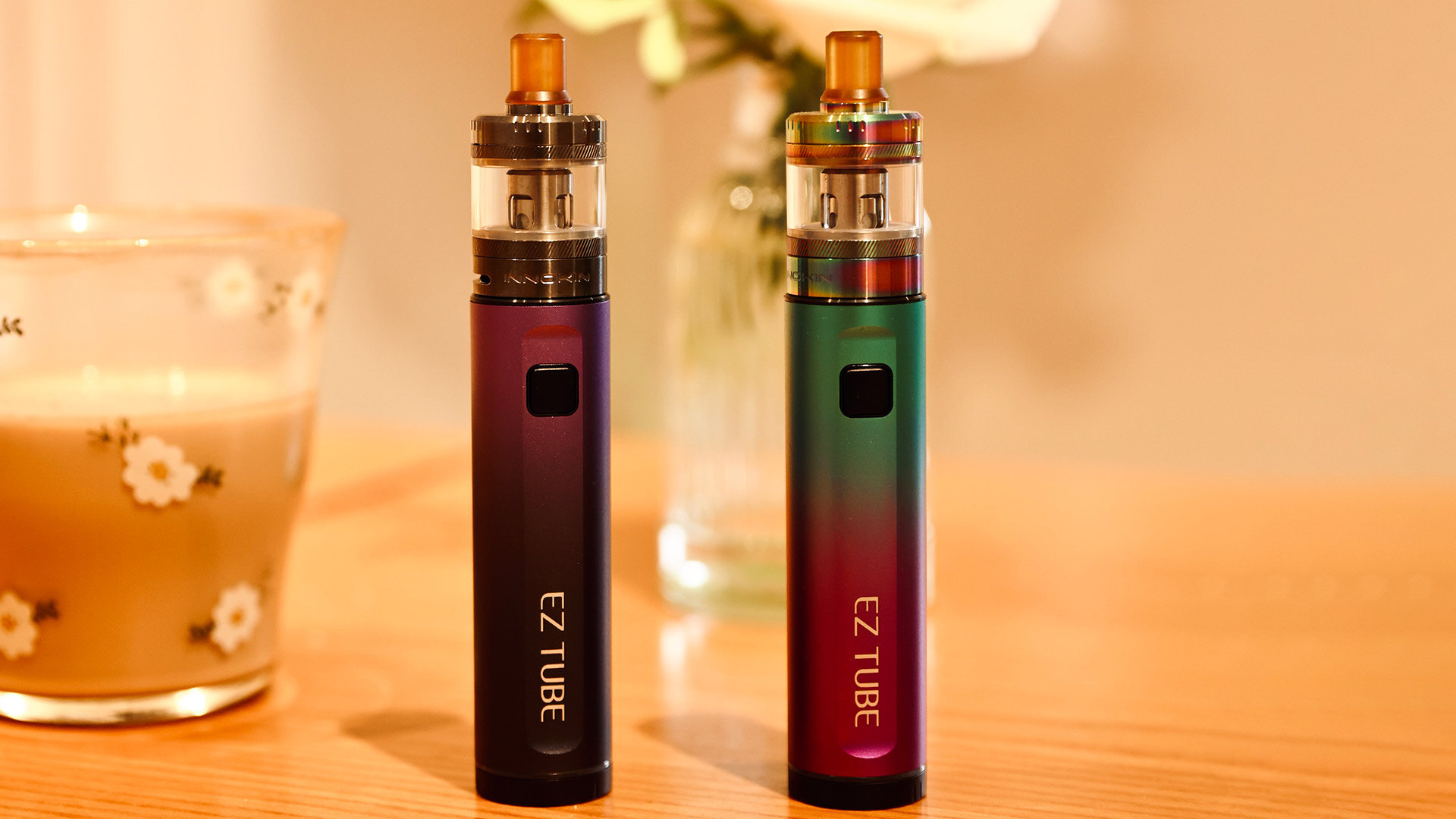 Ein Paar bunte EZ TUBE Vape Pens stehen aufrecht auf einem Holztisch neben einem Glas mit Blumenmuster und weisen auf ein häufiges Problem hin - warum ein Vape Pen nach dem Aufladen nicht mehr funktioniert, z. B. weil sich der Akku des Geräts dem Ende seiner Lebensdauer nähert.