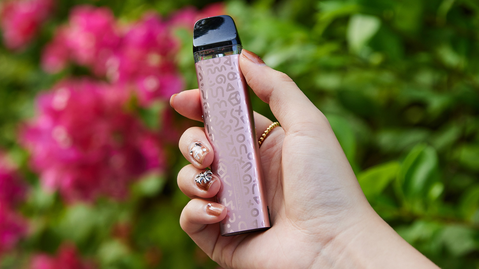 Eine Hand hält den Innokin Trine Pod Vape in Roségold, verziert mit einem dezenten Muster, vor dem Hintergrund leuchtend pinkfarbener Blumen.