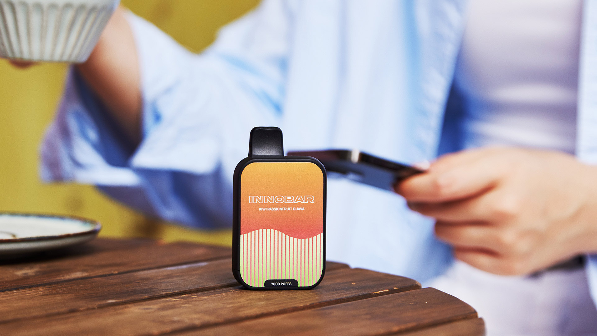 Ein Einweg-Vape-Gerät mit der Aufschrift INNOBAR und 7000 Puffs auf einer hölzernen Oberfläche, mit einem Mädchen, das eine Tasse Kaffee hebt, was auf ein dauerhaftes und befriedigendes Dampferlebnis hindeutet.